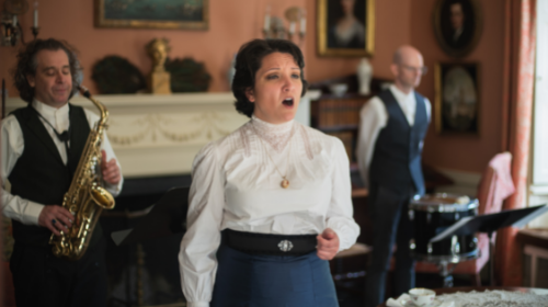 Woman singing in the parlor with two musicians in background.