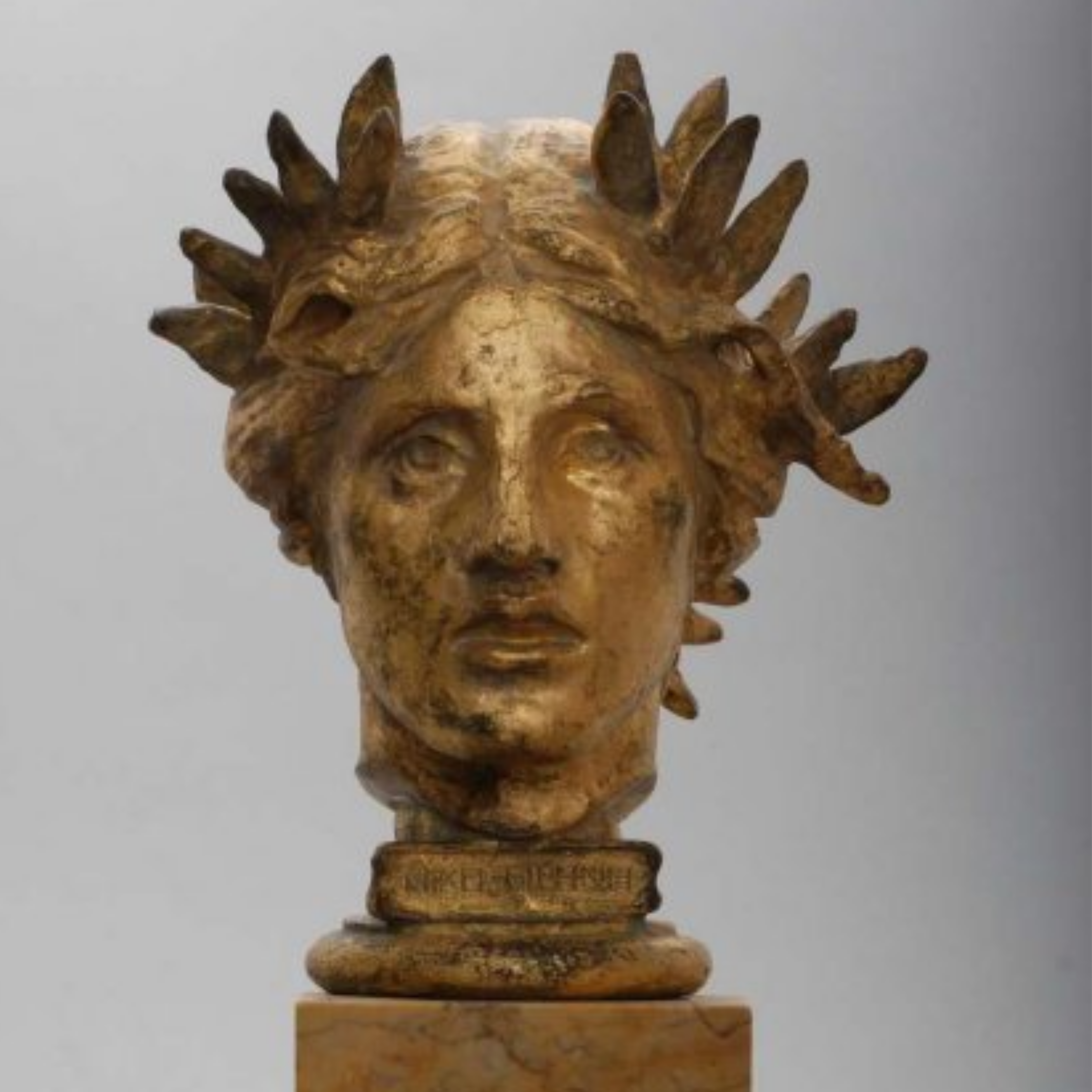 Gilded bronze bust of Victory with a garland on her head, gray background.